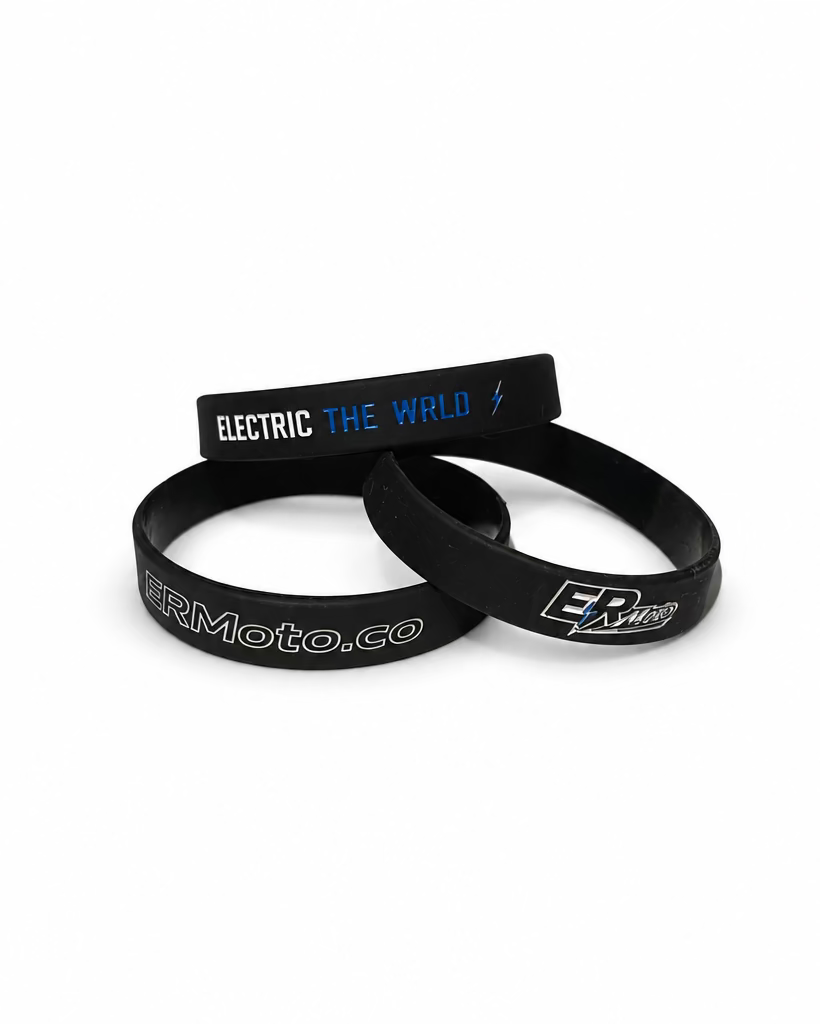 Three black silicone wristbands with white and blue printed graphics, including 'ELECTRIC THE WORLD' and 'Moto.co', displayed on a light-colored tabletop near a window.